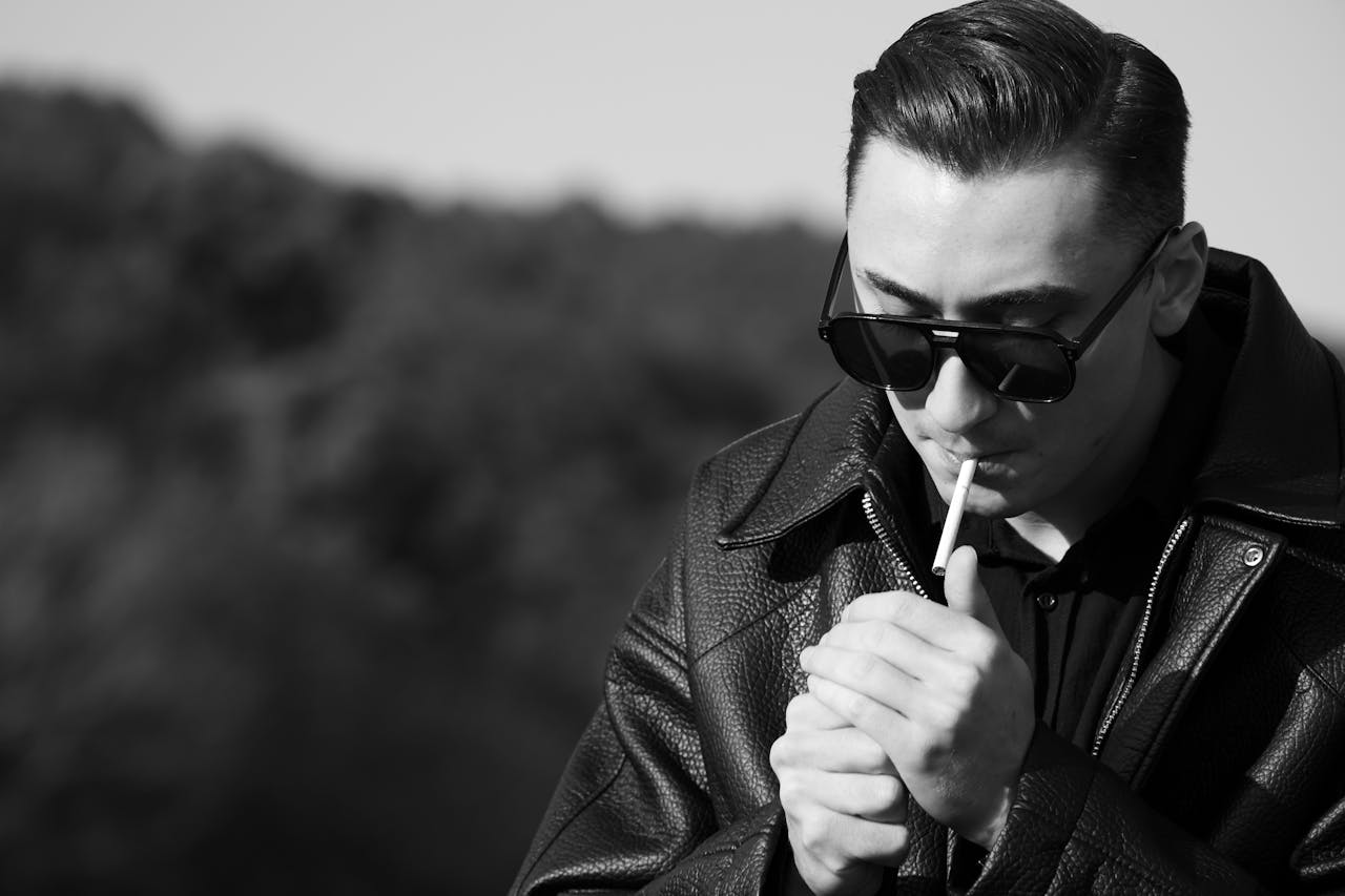 A fashionable man in sunglasses and leather jacket lighting a cigarette outdoors in Istanbul, Türkiye.